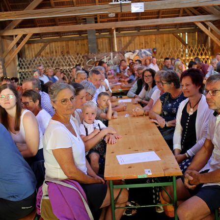 Eine Gruppe von Menschen, einschließlich Erwachsener und Kindern, sitzen in einem Holzsaal. Sie sitzen an langen Tischen, einige mit Papieren, wahrscheinlich während einer Gemeinschaftsveranstaltung.