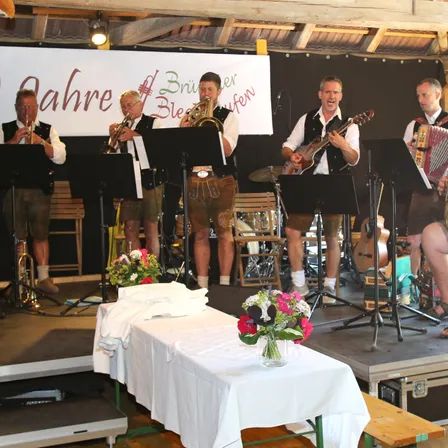 Fünf Musiker spielen auf der Bühne und tragen traditionelle bayerische Kleidung. Einer spielt Gitarre, ein anderer Trompete und andere Akkordeon und Schlagzeug. Vor ihnen steht ein Tisch mit Blumen.