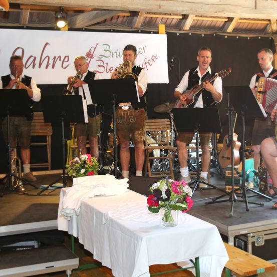 Fünf Musiker spielen auf der Bühne und tragen traditionelle bayerische Kleidung. Einer spielt Gitarre, ein anderer Trompete und andere Akkordeon und Schlagzeug. Vor ihnen steht ein Tisch mit Blumen.