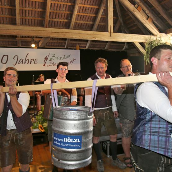 Eine Gruppe von Männern in traditioneller bayerischer Tracht hebt ein Fass auf einem Balken, während zwei Männer mit Mikrofonen hinter ihnen stehen, und ein Schild steht '10 Jahre Brindler Bleufern.'