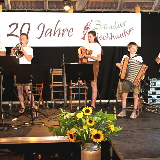 Eine Band spielt auf der Bühne mit einem Banner, das '20 Jahre Brindler Lechhausen' besagt. Einer spielt Trompete, ein anderer Gitarre, und ein Junge spielt Akkordeon.