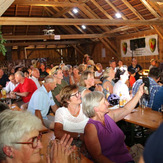 Eine Gruppe von Menschen, überwiegend ältere Erwachsene, sitzt um Tische herum in einer Scheune. Einige klatschen, und eine Frau macht ein Foto mit ihrem Handy. An den Wänden hängen Banner.