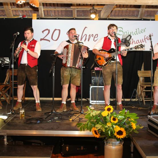 Vier Musiker spielen auf der Bühne, zwei mit Instrumenten in den Händen, einer spielt eine Gitarre, der andere eine Zieharmonica. Hinter ihnen steht ein Banner mit der Nummer 20.