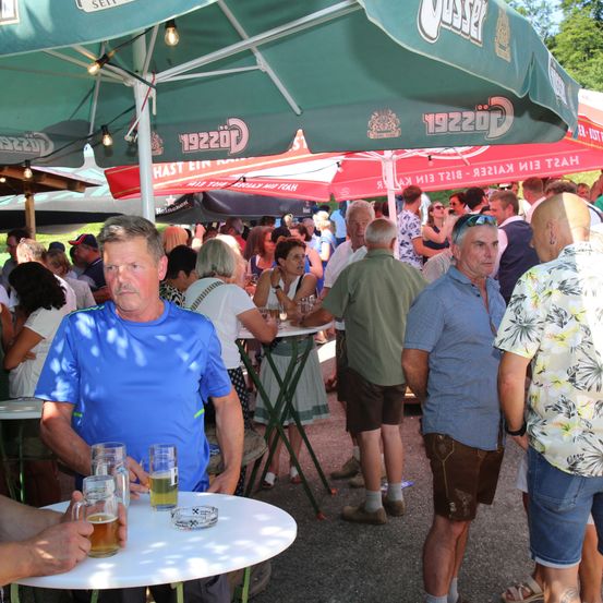 Eine Outdoor-Versammlung mit Menschen unter Regenschirmen. Einige sitzen und trinken Bier, während andere stehen. Die Umgebung ist lässig, mit Tischen und Stühlen. Es gibt grüne Regenschirme mit Text und Logos.