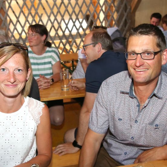 Eine Gruppe von Menschen sitzt an einem Tisch in einem Restaurant. Eine Frau und ein Mann lächeln in die Kamera. Hinter ihnen unterhalten sich ein weiterer Mann und eine Frau. Der Mann trägt eine Brille.