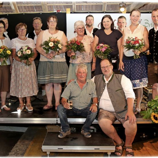Eine Gruppe von Menschen steht auf einer Bühne, hält Blumen und lächelt für ein Foto. Einige tragen Brillen, und ein Mann hält ein Glas. Hinter ihnen ist ein Banner.