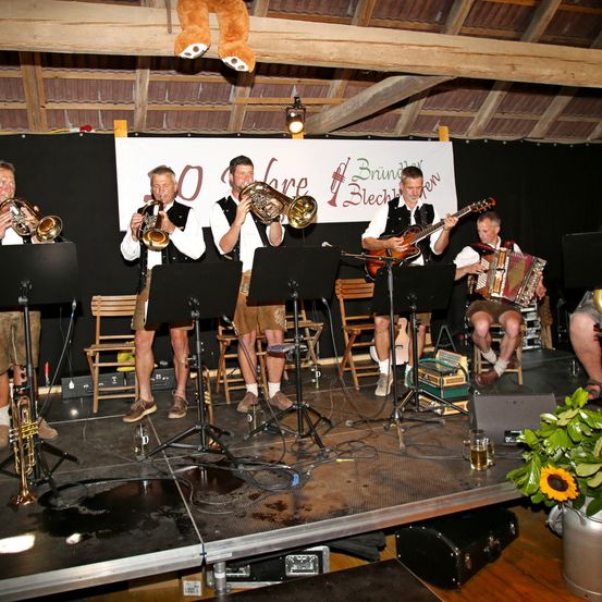 Eine Gruppe von Musikern spielt auf einer Bühne mit einem Banner hinter ihnen, das '30 Jahre' steht. Sie spielen verschiedene Instrumente, darunter Trompeten, Posaunen, eine Gitarre und ein Akkordeon. Die Bühne ist mit Stühlen, Mikrofonen und einem Lautsprecher ausgestattet.