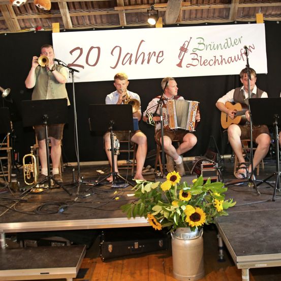 Fünf Männer spielen auf einer Bühne mit Musikinstrumenten, ein Banner hinter ihnen lautet '20 Jahre Bründler Blechhauvon'.