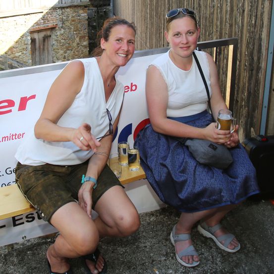 Zwei Frauen sitzen auf einer Bank, eine mit einem Getränk und Sonnenbrille auf dem Kopf, die andere mit einem Getränk und einer Tasche.
