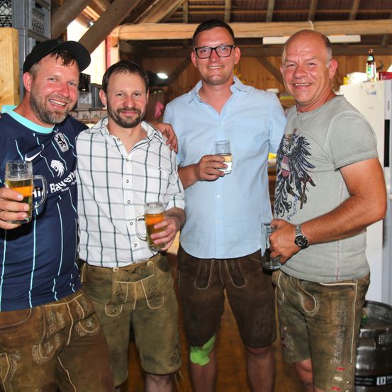 Vier Männer in traditioneller bayerischer Kleidung, sogenannte Lederhosen, posieren für ein Foto, während sie Bierkrüge und Gläser halten. Der Hintergrund scheint ein rustikaler Raum mit Holzwänden zu sein.