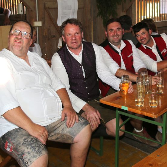 Vier Männer sitzen an einem Tisch mit Gläsern Bier. Der Mann auf der linken Seite trägt eine Brille und ein weißes Hemd. Die anderen tragen rote Westen.