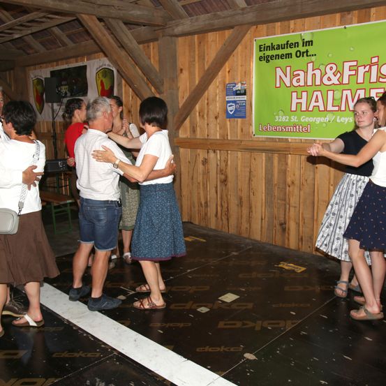 Eine Gruppe von Erwachsenen tanzt in einem hölzernen Raum mit einem grünen Banner, das Nah&Frisch HALM liest. Sie sind zu zweit und halten sich an den Armen. Der Boden hat weiße Linien.