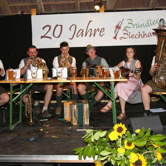 Eine Gruppe Musiker tritt auf der Bühne auf, gekennzeichnet durch ein Banner, das 20 Jahre 'Bründlär Blechhaus' feiert. Sie spielen Instrumente wie Trompeten, Saxophone und Gitarren. Vier Personen sitzen auf Bänken, mit Biergläsern auf dem Tisch, und eine Tuba rechts. Vorne stehen Sonnenblumen und grüne Blätter.