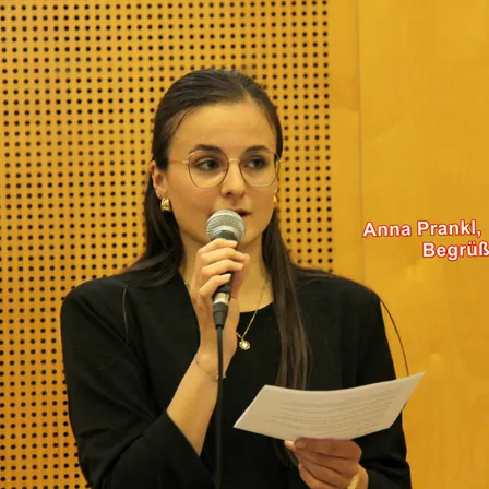 Anna Prankl hält ein Mikrofon und ein Stück Papier in der Hand und steht vor einer gemusterten Wand. Sie trägt eine Brille und eine Halskette.