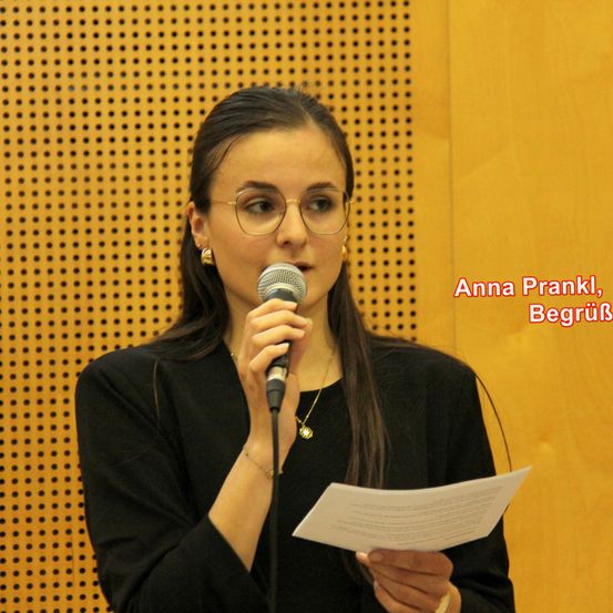 Anna Prankl hält ein Mikrofon und ein Stück Papier in der Hand und steht vor einer gemusterten Wand. Sie trägt eine Brille und eine Halskette.