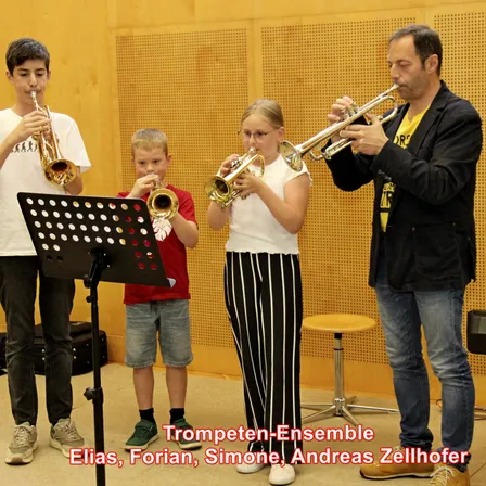 Drei Kinder und ein Mann spielen Trompeten in einem Studio mit einem Notenständer und einer Holzwand. Der Junge links trägt ein weißes T-Shirt, der Junge in der Mitte ein rotes T-Shirt und das Mädchen ein weißes Top mit schwarzer Hose. Der Mann trägt einen schwarzen Anzug und Jeans.