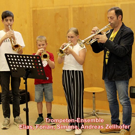 Drei Kinder und ein Mann spielen Trompeten in einem Studio mit einem Notenständer und einer Holzwand. Der Junge links trägt ein weißes T-Shirt, der Junge in der Mitte ein rotes T-Shirt und das Mädchen ein weißes Top mit schwarzer Hose. Der Mann trägt einen schwarzen Anzug und Jeans.