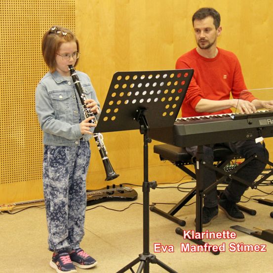 Ein junges Mädchen steht in einem Klassenzimmer und spielt Klarinette, während ein Mann neben ihr das Klavier spielt. Das Mädchen trägt eine Brille und eine Jeansjacke. Die Szene umfasst einen Notenständer mit Musiknoten, ein Keyboard und eine Gitarre im Hintergrund.