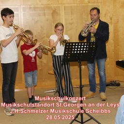 Vier Personen, darunter zwei Kinder, spielen Trompeten in einer Musikschule. Ein Notenständer steht vor ihnen. Das Datum 28.05.2025 wird angezeigt.