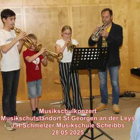 Vier Personen, darunter zwei Kinder, spielen Trompeten in einer Musikschule. Ein Notenständer steht vor ihnen. Das Datum 28.05.2025 wird angezeigt.