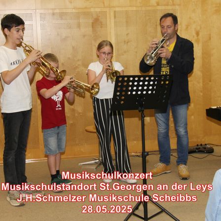 Vier Personen, darunter zwei Kinder, spielen Trompeten in einer Musikschule. Ein Notenständer steht vor ihnen. Das Datum 28.05.2025 wird angezeigt.