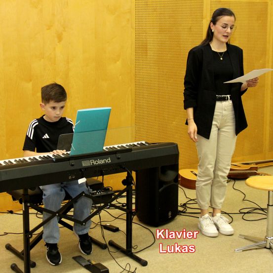 Ein Junge spielt Klavier, während eine Frau neben ihm steht und ein Blatt Papier in der Hand hält. Auf dem Klavier steht der Name 'Roland'. Der Junge trägt ein schwarzes T-Shirt und Jeans. Die Frau trägt einen schwarzen Blazer, weiße Hosen und weiße Turnschuhe.