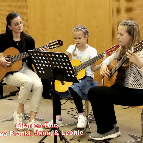 Drei Personen sitzen auf Hockern und spielen Gitarre. Die Frau links trägt eine Brille und eine Halskette. Die Mittlere ist ein junges Mädchen mit Stirnband. Das Mädchen rechts trägt eine Kleidung mit Punkten. Der Text lautet 'Gitarren-Duo Anna Prankl, Jana & Leonie.'