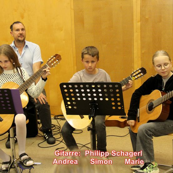 Vier Kinder spielen Gitarre in einem Raum, zwei Jungen und zwei Mädchen. Sie haben einen Notenständer vor sich mit Noten. Ein erwachsener Mann steht hinter ihnen. Die Namen der Kinder sind in rotem Text geschrieben.