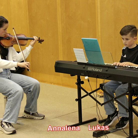 Zwei junge Musiker, Annalena und Lukas, spielen ein Duett auf Klavier und Geige. Annalena sitzt und hält eine Geige, während Lukas auf einem Stuhl sitzt und Klavier spielt. Beide tragen Freizeitkleidung.