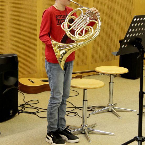 Ein junger Junge steht in einem Raum, hält eine französische Horn, mit einer Gitarre, Lautsprecher und Notenständern im Hintergrund.