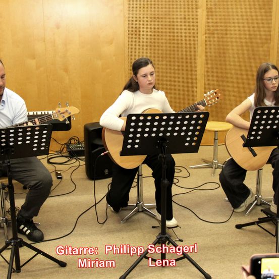 Drei Personen spielen Gitarren in einem Raum. Die mittlere Person, ein Mädchen, spielt eine klassische Gitarre. Ein Mann links und ein Mädchen rechts spielen E-Gitarren. Der Raum hat eine Holzwand.