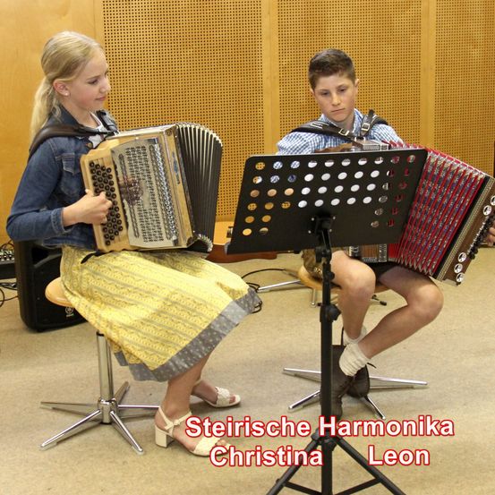 Zwei junge Musiker spielen Akkordeon. Das Mädchen, Christina, trägt einen gelben Rock. Der Junge, Leon, trägt ein blaues Hemd. Hinter ihnen ist eine Holzwand und ein Lautsprecher.