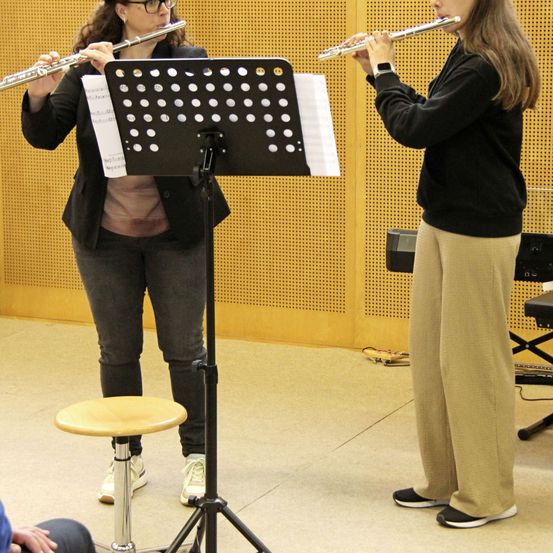 Zwei Frauen spielen Flöten in einem Musikraum, eine steht, eine sitzt, mit einem Notenständer dazwischen.