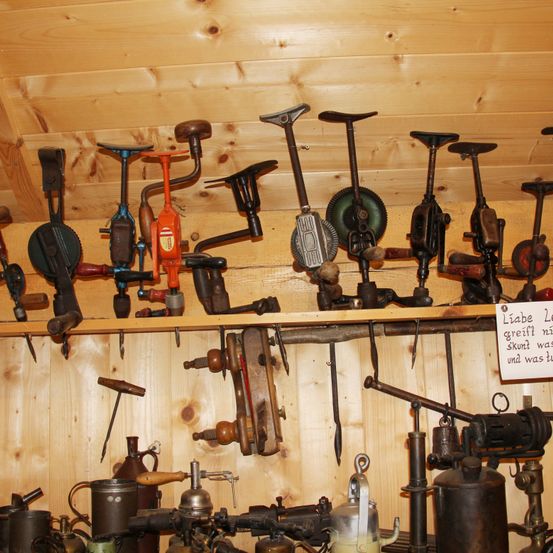 Ein hölzernes Regal hält verschiedene altmodische Werkzeuge. Einige sind rostig, andere haben Holzgriffe. Ein Schild an der Wand lautet 'Liebe Legriff mit'.