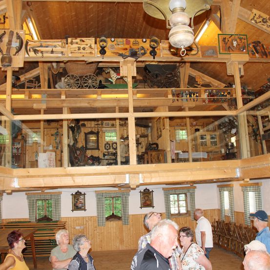 Eine Gruppe älterer Menschen besucht ein Museum. Der Innenraum ist holzverkleidet mit verschiedenen historischen Gegenständen. Es stehen Menschen und schauen nach oben zur hölzernen Decke.