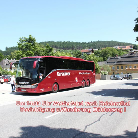 Ein roter Bus steht am Straßenrand mit einem Text in Deutsch darüber. Ein Mann steht neben dem Bus. Gebäude und Autos sind am Straßenrand geparkt. Bäume und Berge sind in der Ferne.