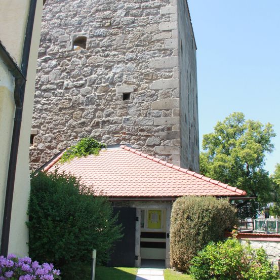 Ein hoher Steinturm mit einem roten Dach und einem kleinen angebautem Gebäude, umgeben von grünen Pflanzen und Blumen, unter einem klaren blauen Himmel.