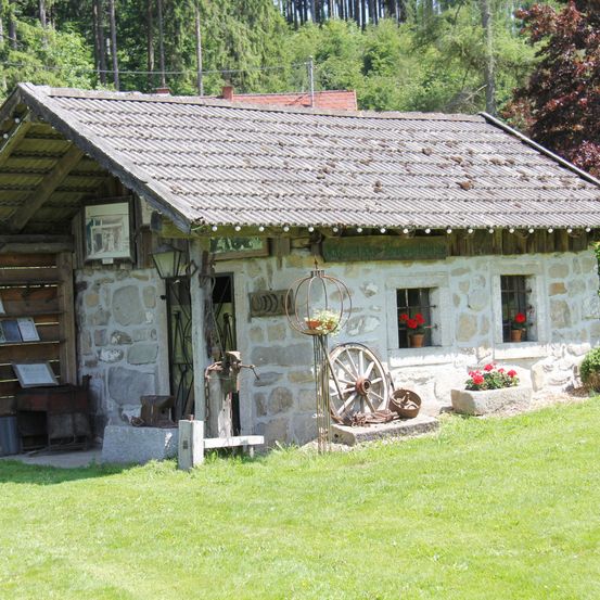 Ein kleines Steinhaus mit einem Metalldach, in einem üppigen grünen Feld gelegen. Es hat ein Rad und eine Lampe draußen und zwei Fenster mit roten Blumen.