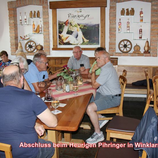 Mehrere Erwachsene sitzen um einen Tisch in einem Restaurant. Sie haben Weingläser, eine Flasche und eine Blume auf dem Tisch. Das Bild hat einen Wasserzeichen mit dem Text 'Abschluss beim Heurigen Pihringer in Winkl'.