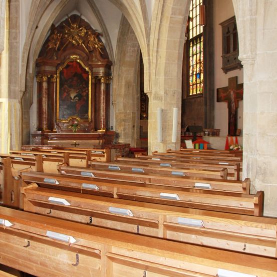 Das Innere einer Kirche zeigt Holzbänke, einen großen Altar und Buntglasfenster. Der Altar hat einen goldenen Rahmen und ein Gemälde. Ein Kreuz ist an der Wand befestigt.