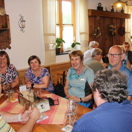 Eine Gruppe von Menschen sitzt in einem gemütlichen, rustikalen Restaurant an einem Tisch und lächelt und plaudert. Der Tisch ist mit Gläsern, einem gerahmten Foto und einem Kerzenständer gedeckt. Im Hintergrund befinden sich Holzwände und ein Fenster mit einer Pflanze.