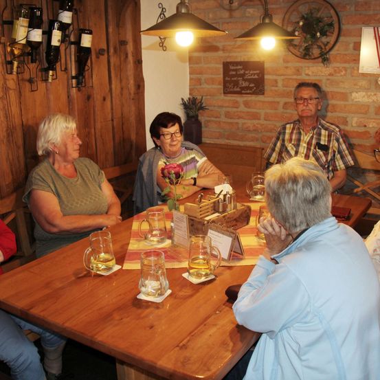 Eine Gruppe älterer Personen sitzt um einen Holztisch in einem gemütlichen, rustikalen Raum. Der Tisch ist mit Gläsern und einer Rose als Mittelpunkt geschmückt. Der Raum hat Holzwände, hängende Lampen und eine dekorative Ziegelwand.