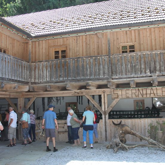 Eine Gruppe von Menschen steht in einem Holzgebäude, möglicherweise einem Museum. Sie betrachten ausgestellte Gegenstände und das Wort "STERNTALE" ist an der Wand. Draußen befinden sich Baumstämme und Pflanzen.