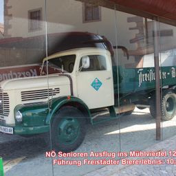 Ein alter weiß-grüner Lastwagen wird in einer Glasvitrine ausgestellt, mit Texten auf Deutsch. Der Lastwagen steht auf einer Kopfsteinpflasterstraße.