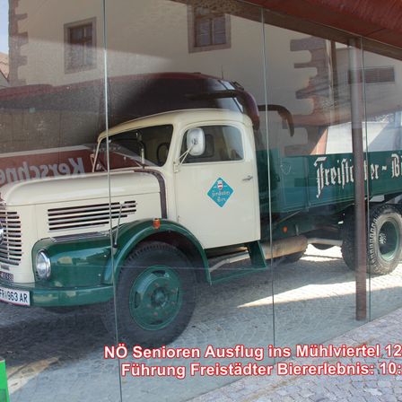 Ein alter weiß-grüner Lastwagen wird in einer Glasvitrine ausgestellt, mit Texten auf Deutsch. Der Lastwagen steht auf einer Kopfsteinpflasterstraße.