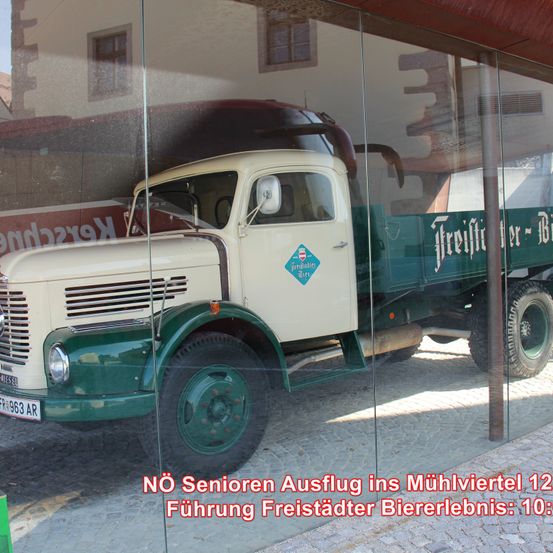 Ein alter weiß-grüner Lastwagen wird in einer Glasvitrine ausgestellt, mit Texten auf Deutsch. Der Lastwagen steht auf einer Kopfsteinpflasterstraße.