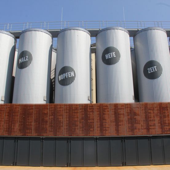Eine Reihe von vier großen silbernen Metalltanks, jeweils mit einem Wort beschriftet: Malz, Hopfen, Hefe und Zeit. Sie stehen auf einer Metallplattform unter einem blauen Himmel.
