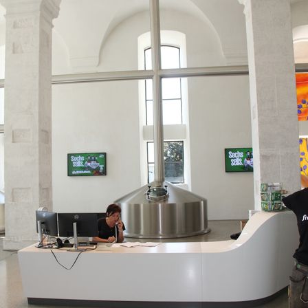 Eine Frau arbeitet an einer Rezeption mit Monitoren. Hinter ihr ist ein großer Edelstahltank zu sehen. Zwei Monitore zeigen Werbung an. Die Wände sind weiß und es gibt bogenförmige Fenster mit bogenförmigen Rahmen.