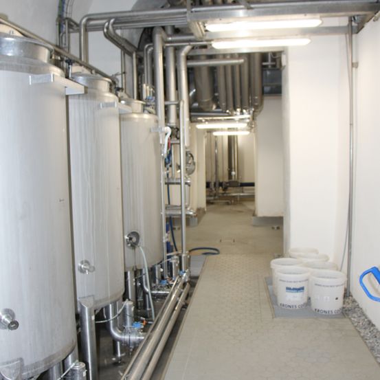 Ein industrielles Umfeld mit großen Edelstahltanks, die durch Rohre verbunden sind. Weiße Eimer sind an einer Wand aufgestellt. Der Bereich ist gut beleuchtet durch Deckenleuchten.