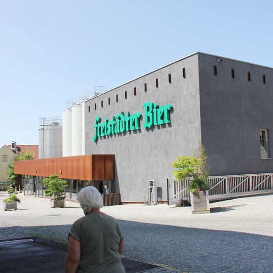 Eine ältere Frau geht vor dem Freistadter Bier Gebäude mit grüner Beschilderung und modernem Architekturdesign. Das Gebäude hat große Fenster und einen braunen Vorbau.
